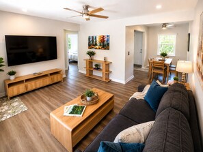 Ein modernes Wohnzimmer mit Flachbildfernseher, Holzmöbeln, Deckenventilator und einem Essbereich mit Tageslicht.