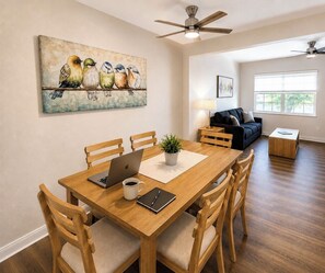 Ein Essbereich mit Holztisch, Stühlen, einem Laptop, einer Kaffeetasse und einer Pflanze. An der Wand hängt ein Vogelgemälde, es gibt einen Deckenventilator und im Hintergrund eine Couch.