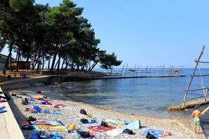 Een strand met parasols en handdoeken op het kiezelstrand, een houten pier die het water in loopt, en bomen langs de kust.