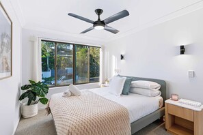 A bedroom with a bed, a ceiling fan, a nightstand, a potted plant, and a window with a view of greenery. - Chic Noosa Pad - Walk to Noosa Hastings St (Noosa)