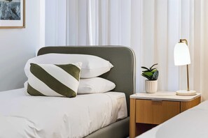 Ein ordentlich bezogenes Bett mit weiß-grünen Kissen, einer Zimmerpflanze auf dem Nachttisch und einer Nachttischlampe.