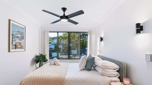 A bedroom with a bed, a bedside table, a nightstand, a ceiling fan, a window with curtains, a wall-mounted light, and a framed picture. - Chic Noosa Pad - Walk to Noosa Hastings St (Noosa)