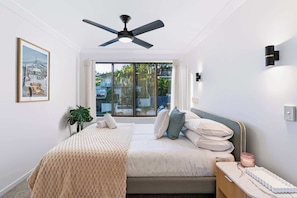 Ein Schlafzimmer mit Bett, Nachttisch, Deckenventilator, Fenster mit Vorhängen, Wandleuchte und einem gerahmten Bild.