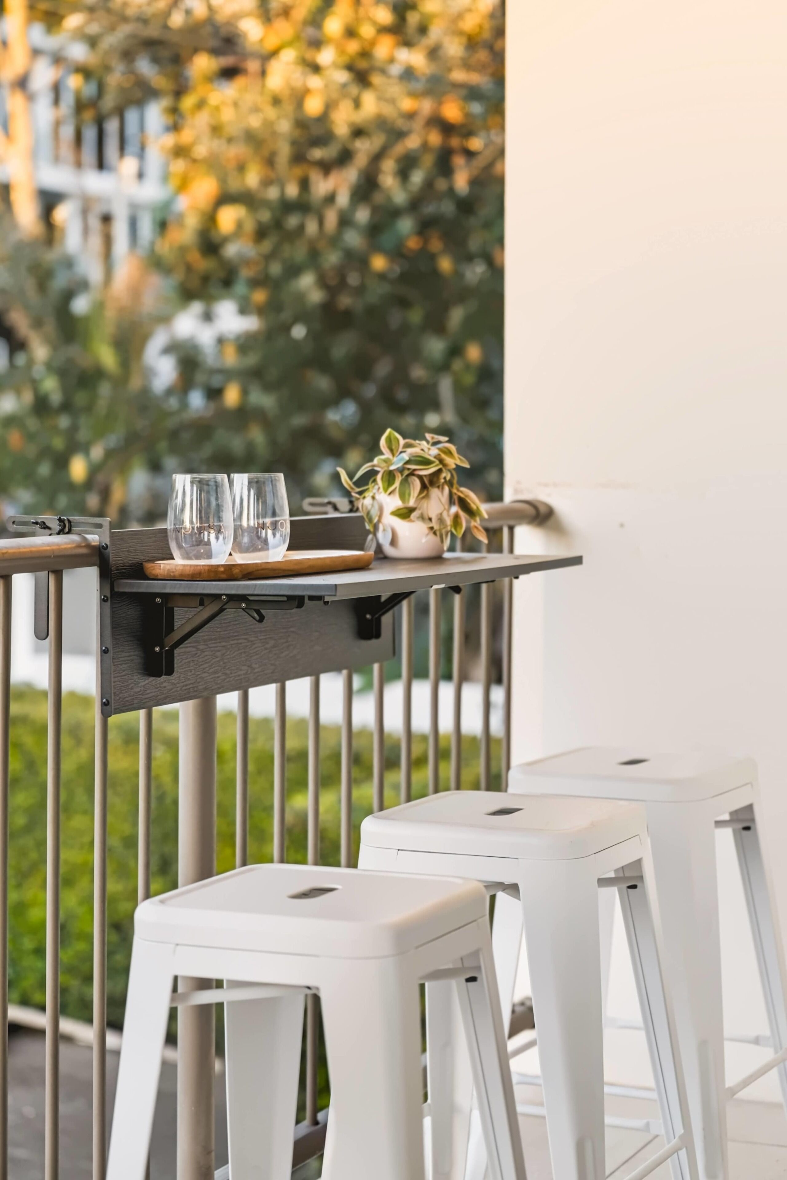 A balcony with a table set for two, featuring two white stools and a small potted plant.