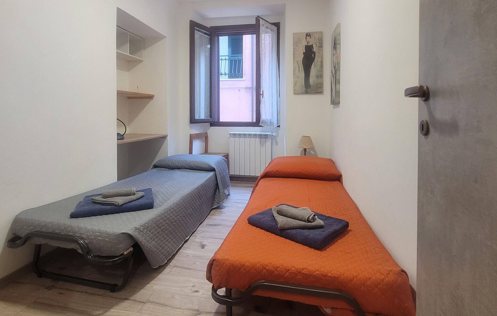 A small, narrow room with two single beds, a radiator, a window with a view, and a door.