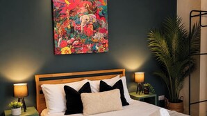 A neatly made bed with a patterned bedspread and pillows, flanked by two bedside tables with lamps and books. A colorful abstract painting adorns the wall above the bed. - 3BR Gateshead Home | Free Parking & WiFi (Gateshead)