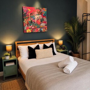 Uma cama bem arrumada com colcha e almofadas estampadas, com duas mesas de cabeceira laterais com luminárias e livros. Um quadro abstrato colorido decora a parede acima da cama.