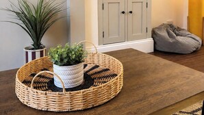 A living room with a television mounted on the wall, a bookshelf with books and decorative items, a grey bean bag chair, and a round woven table with a potted plant. - 3BR Gateshead Home | Free Parking & WiFi (Gateshead)