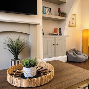 Uma sala de estar com uma televisão na parede, uma estante com livros e objetos decorativos, uma poltrona de couro flexível cinza e uma mesa redonda de fibra natural com um vaso de planta.