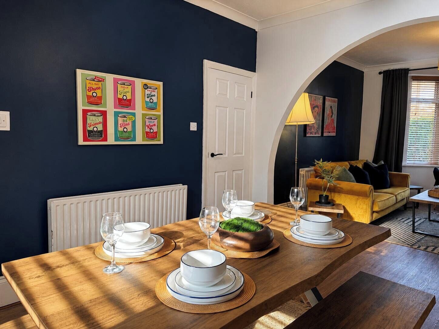 A dining area with a wooden table set for four, a white door, and a dark blue wall with vintage can posters.