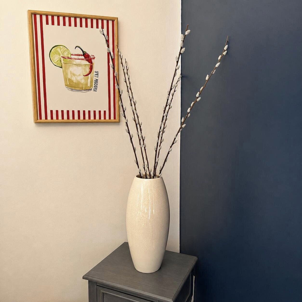 En vit vas med grenar står på ett mörkt bord framför en vägg där ett inramat fotografi hänger.