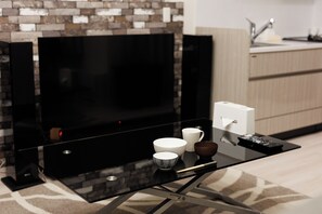 A modern living room with a flat-screen TV mounted on a brick wall, a glass coffee table with remote controls, and a kitchen area in the background. - Mondomio 208 (Sapporo)
