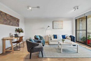 Un salon moderne avec un bureau en bois, deux fauteuils, un canapé et une table basse. Une grande fenêtre offre une vue sur un immeuble en briques.