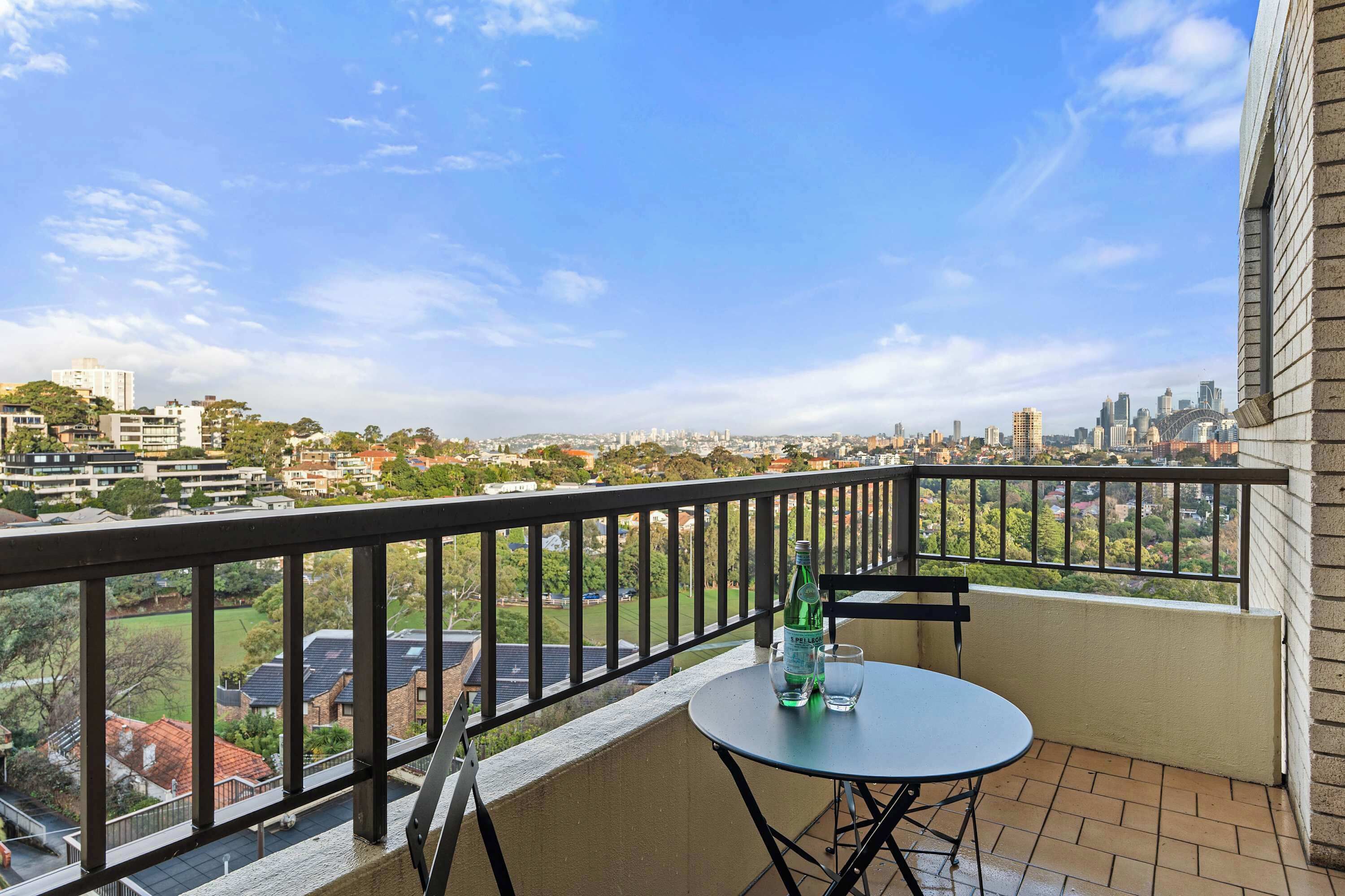A balcony with a table and chairs, overlooking a cityscape with buildings and greenery.