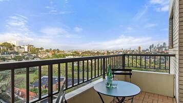 A balcony with a table and chairs, overlooking a cityscape with buildings and greenery.