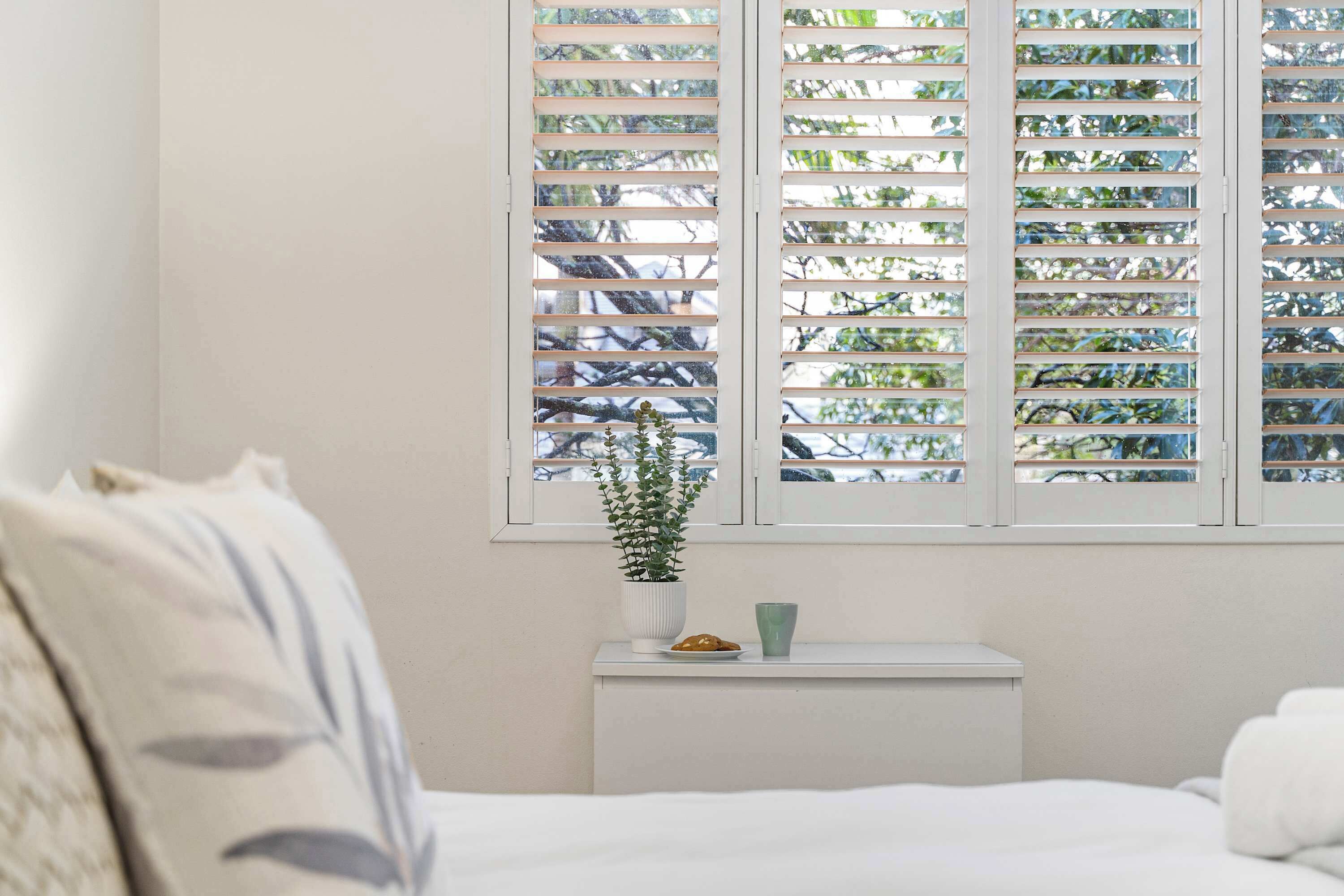 Une chambre à coucher avec un lit, une table de chevet ornée d’une plante et une fenêtre avec des stores.