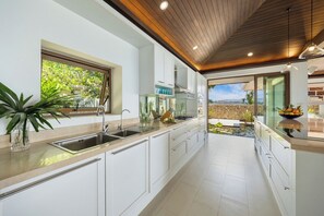 A modern kitchen with white cabinetry, stainless steel appliances, and a view of a garden through the window. - San Suk Villa – 4BR Ocean View Family Tropical Escape (Koh Samui)