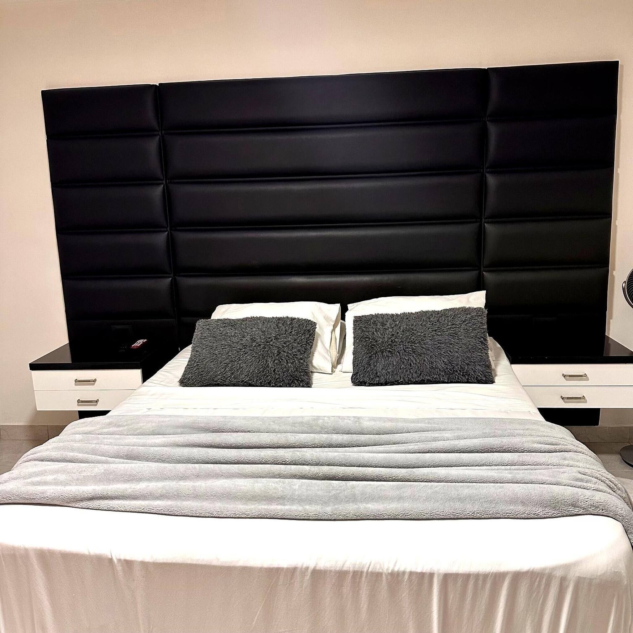 Una cama con cabecera tapizada de color negro, dos almohadas y una mesita de noche blanca con cajones.