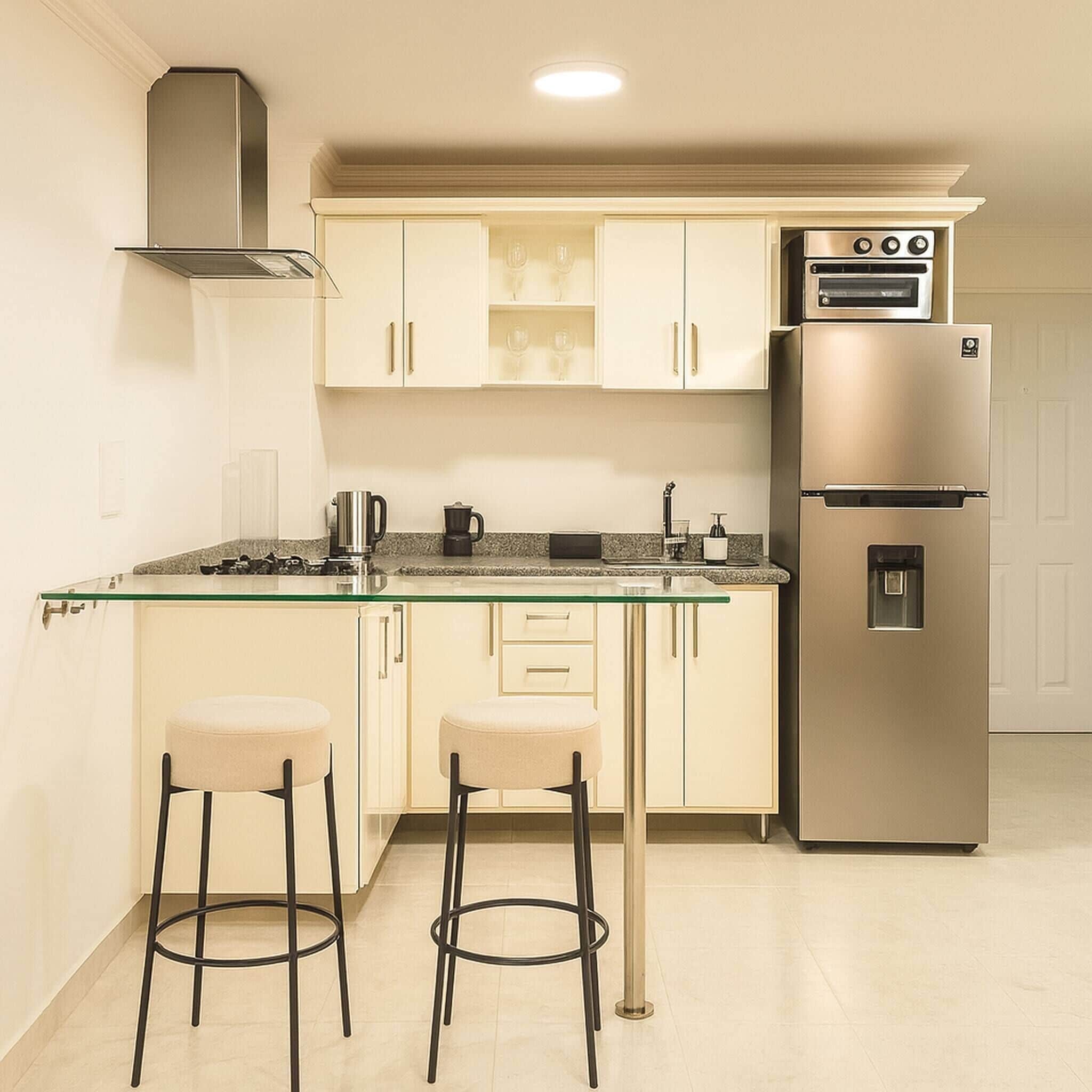 Una cocina compacta con horno empotrado, refrigerador y una isla de vidrio con dos taburetes.