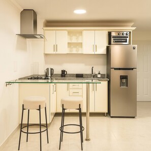 A compact kitchen with a built-in oven, a refrigerator, and a glass-top island with two stools. - Escape Luxury Suite Premium Ultra Fast Wifi (Medellín)