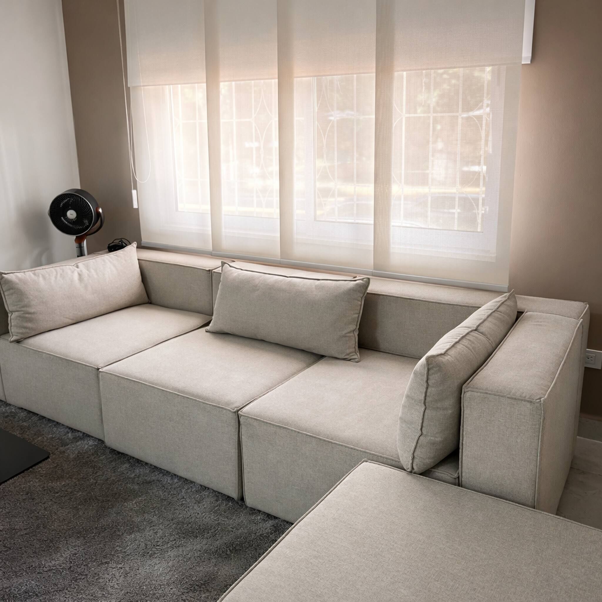 A sectional sofa with a grey fabric, a small black fan, and a window with sheer curtains.
