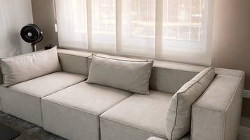 A sectional sofa with a grey fabric, a small black fan, and a window with sheer curtains.
