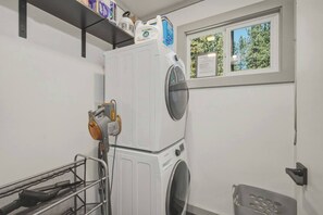 A laundry room with a front-loading washing machine, a dryer, and a shelf above the machine holding cleaning supplies. - Fir Sure Chalet - Dog friendly modern cabin in SLT (South Lake Tahoe)