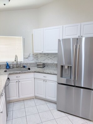 A kitchen with white cabinets, a stainless steel refrigerator, a granite countertop, and a window with blinds. - Modern 3BR Single-Story Home with Spacious Yard (North Las Vegas)