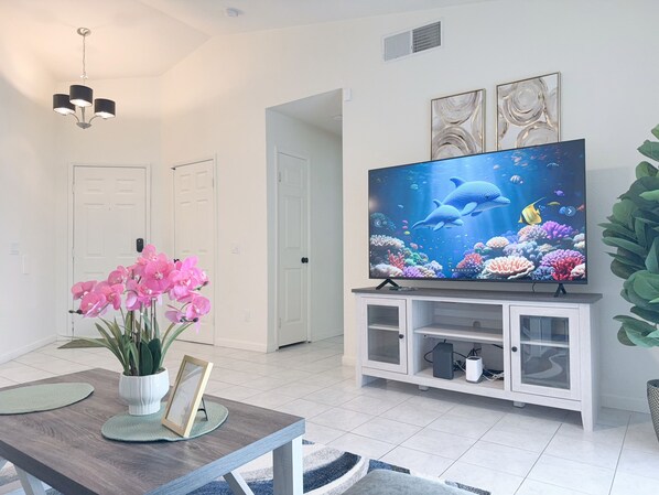 Een woonkamer met een flatscreen televisie, een eettafel met een vaas roze bloemen, en een witte tv-kast.