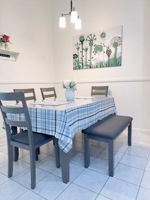 A dining area with a table set for four, featuring a blue and white checkered tablecloth, grey chairs, and a bench. A vase with flowers is on the table, and there is a wall-mounted light fixture above. - Modern 3BR Single-Story Home with Spacious Yard (North Las Vegas)