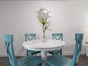 Una mesa redonda blanca con sillas color turquesa, una planta en maceta y un reloj con forma de timón de barco en la pared.