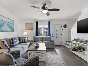 A modern living room with a ceiling fan, a sofa set, a coffee table, and a flat-screen TV. - Coconut Cottage! Golf Cart Friendly Community! (Panama City Beach)