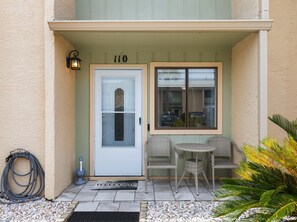 Ein Gebäudeeingang mit einer nummerierten Tür, einer Fußmatte, Sitzgelegenheiten im Freien und einem Gartenbereich.