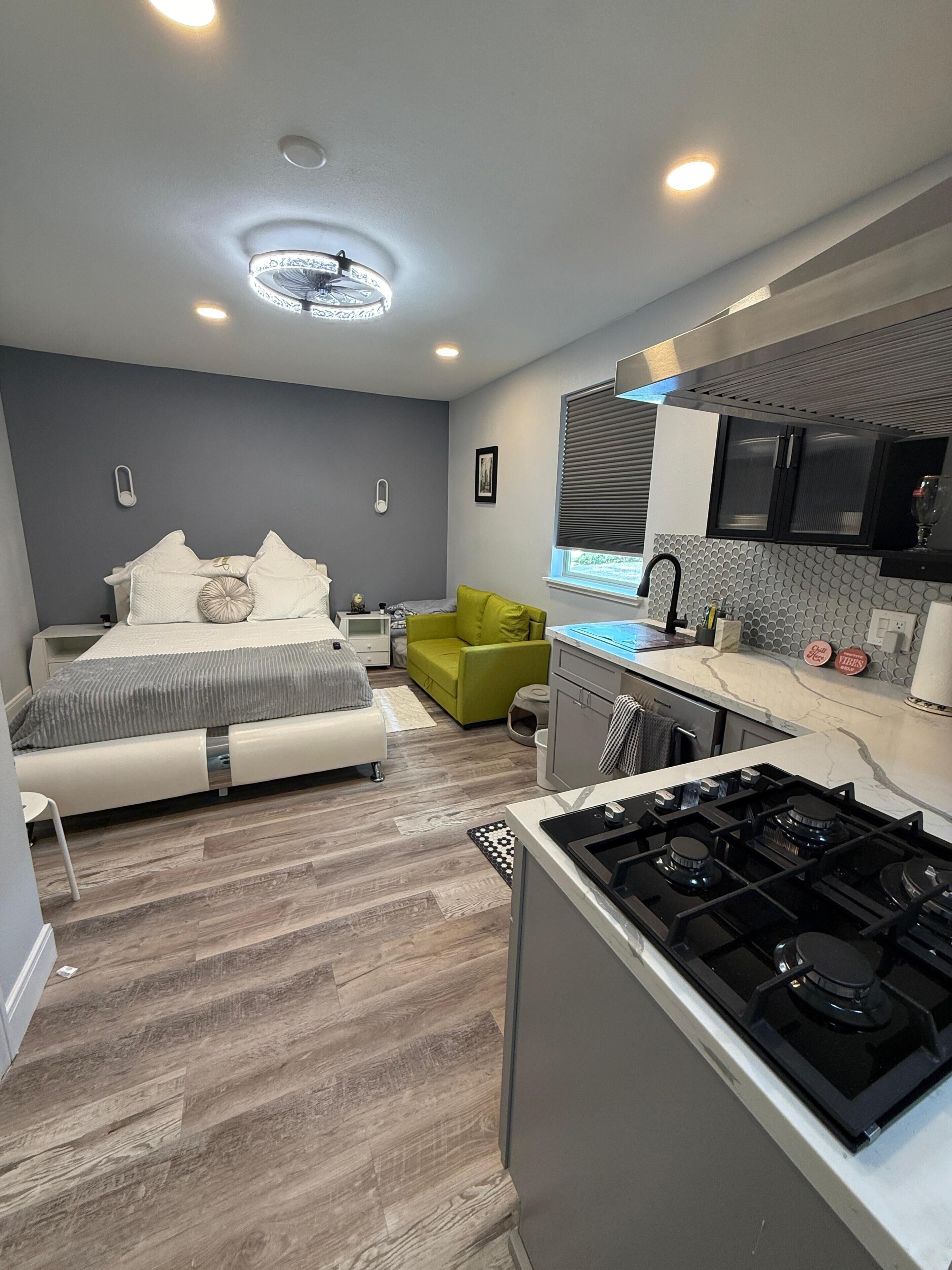 Une chambre à coucher de taille modeste, comprenant un lit, des tables de chevet et un fauteuil vert. À côté se trouve une cuisine moderne équipée d’une cuisinière, d’un évier et d’une fenêtre.