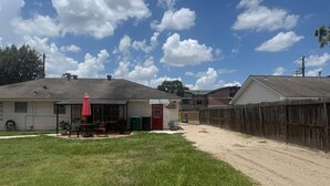 Un barrio residencial con una casa, un patio techado con mobiliario exterior, un jardín cercado y un camino de entrada de grava.