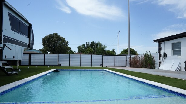 Una piscina con una cerca alrededor, un área verde y un edificio de fachada blanca.