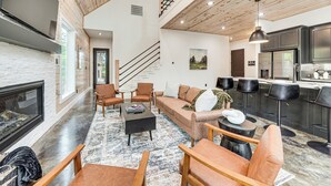Amplia sala de estar con chimenea, asientos cómodos, cocina equipada con electrodomésticos modernos y una escalera que conduce a otro nivel.