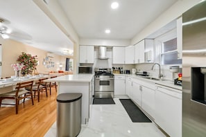 A modern kitchen with white cabinetry, stainless steel appliances, and a dining area with wooden chairs and a marble table. - Charming 3BR Home Near NYC  Free Parking (JERSEY CITY)