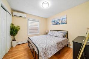 A bedroom with a bed, a potted plant, a window with blinds, a wall-mounted air conditioner, and a painting. - Charming 3BR Home Near NYC  Free Parking (JERSEY CITY)