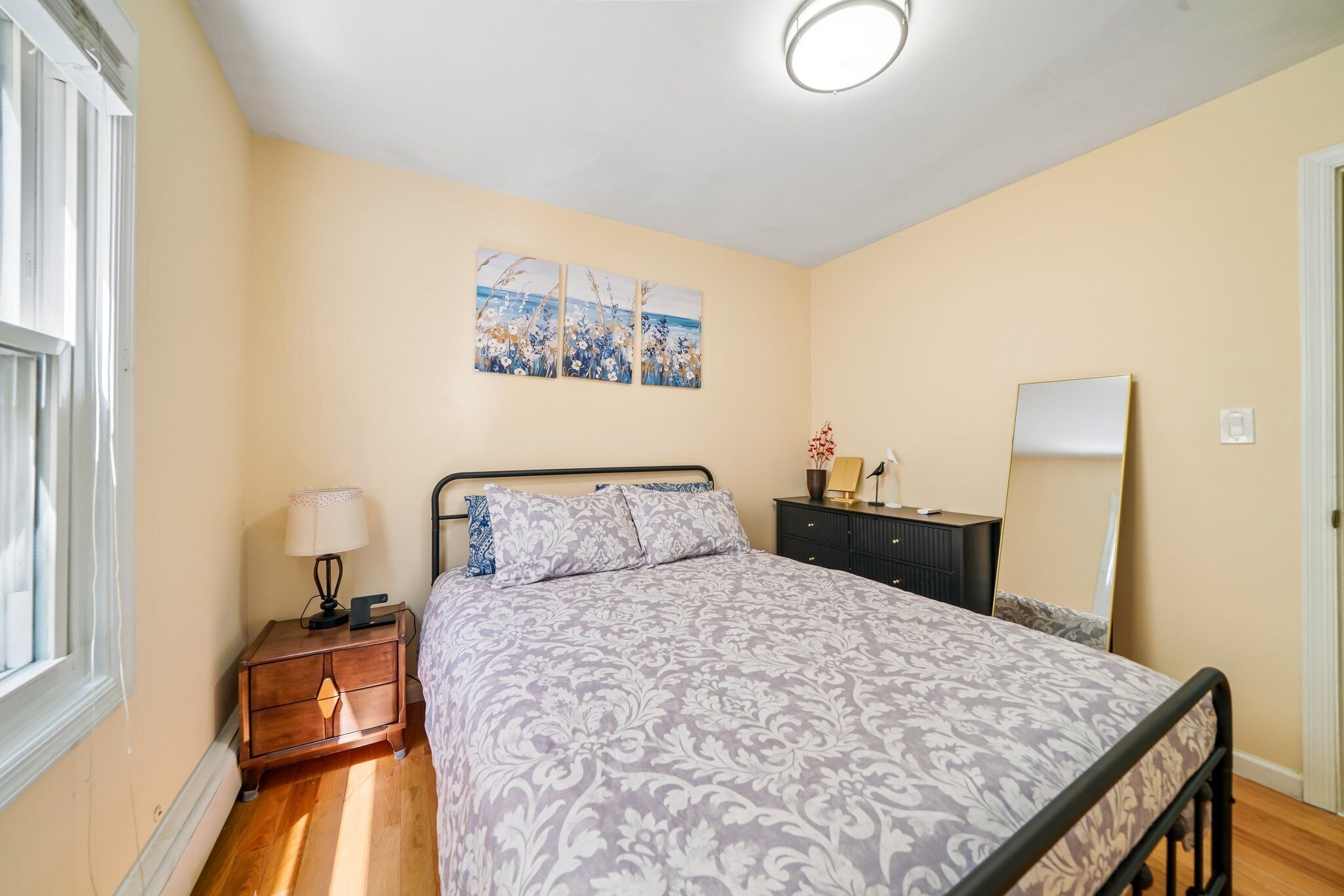 A bedroom with a bed, bedside table, lamp, dresser, and a wall-mounted mirror.