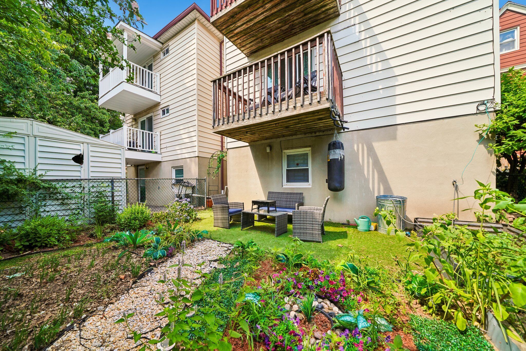 A two-story house with a balcony, a small patio with outdoor furniture, and a garden with various plants and flowers.