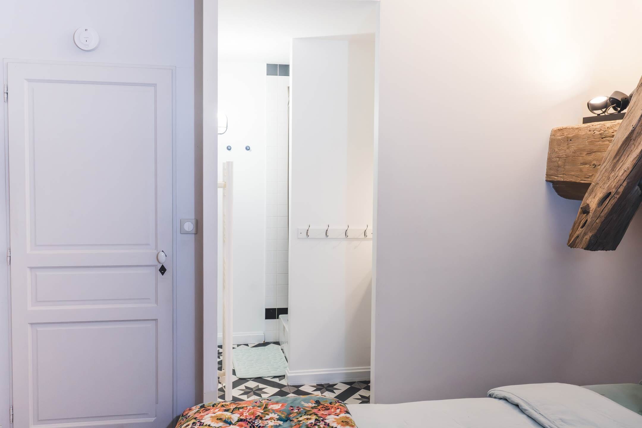 Una habitación con una puerta blanca, una cama con colcha floral, una estantería de madera con una lámpara y un suelo de baldosas.