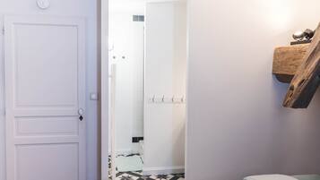 Una habitación con una puerta blanca, una cama con colcha floral, una estantería de madera con una lámpara y un suelo de baldosas.