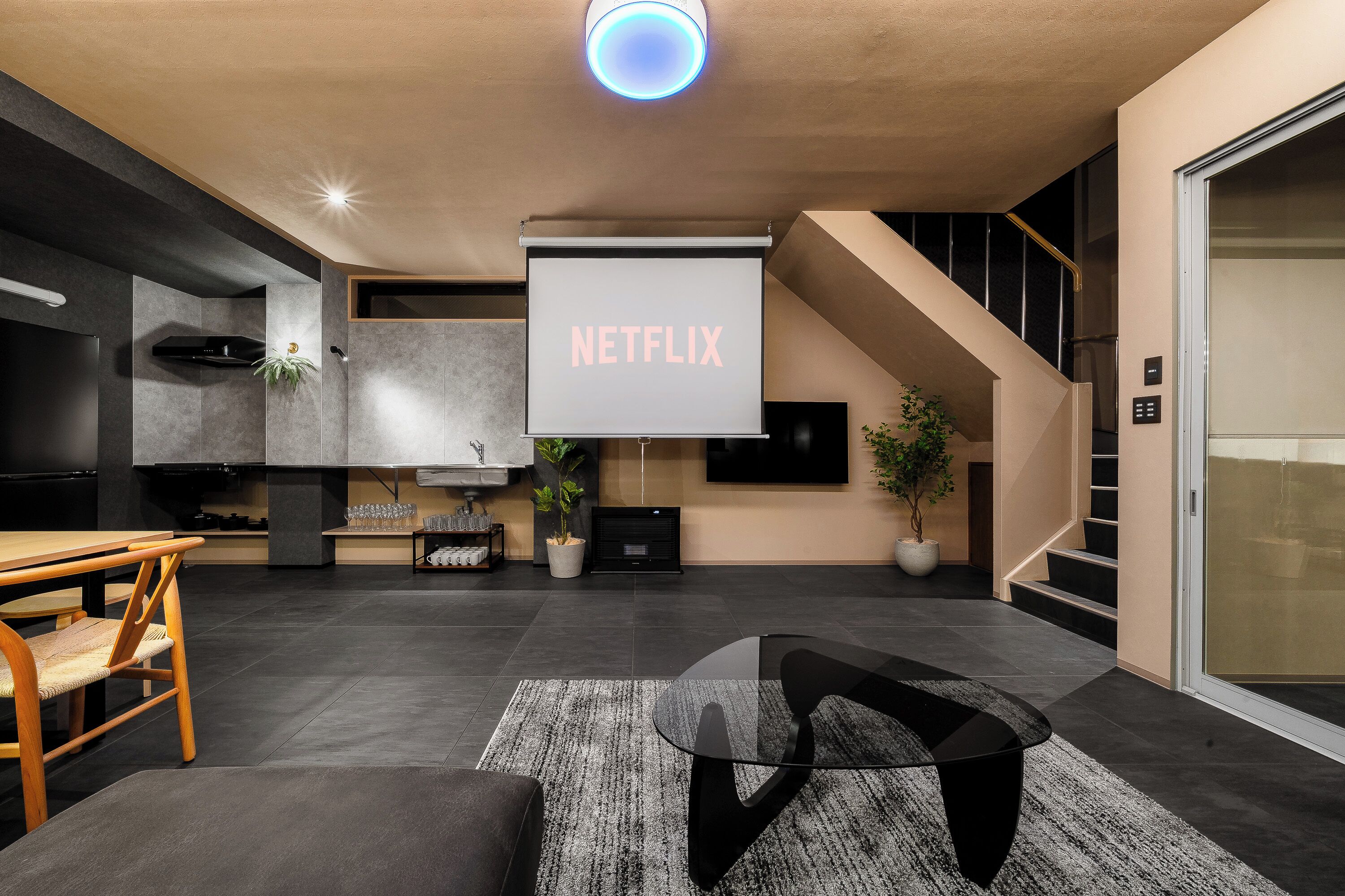 A modern living room with a large screen displaying 'NETFLIX,' a kitchen area, and a staircase.
