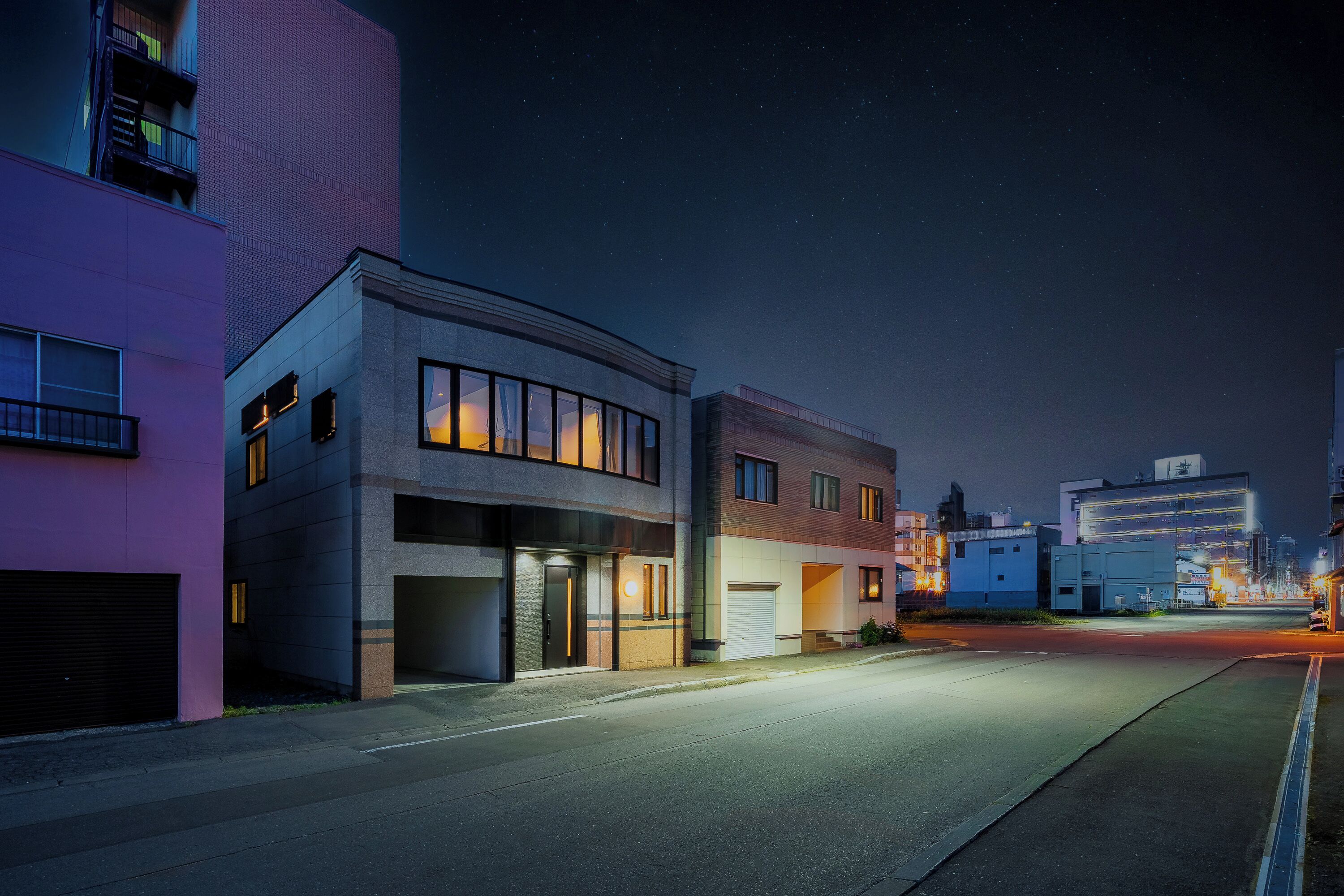 A street at night with modern buildings, a clear sky, and a distant cityscape.