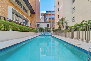 A swimming pool with a tiled edge, surrounded by a low fence, and adjacent to a building with a brick facade and large windows. - Newly Renovated Spacious Apartment (North Sydney)