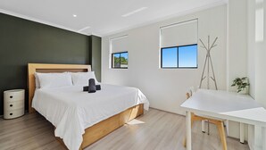 Un dormitorio moderno con una cama de madera, ropa de cama blanca, un escritorio pequeño, una silla y una planta en maceta.