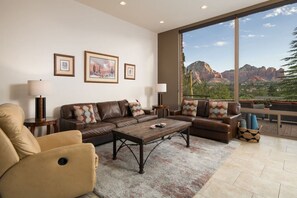 Ein geräumiges Wohnzimmer mit großem Fenster und Blick auf die Berge, ein braunes Ledersofa, ein Couchtisch und ein beigefarbener Sessel.