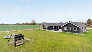 A two-story building with a patio area, a swing set, and a field in the background. - Cozy home in Stege with sauna (Stege)