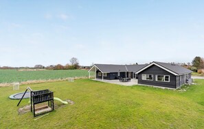 A two-story building with a patio area, a swing set, and a field in the background. - Cozy home in Stege with sauna (Stege)